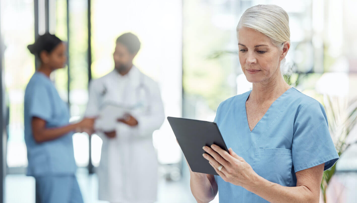 Pflegekraft mit Tablet, im Hintergrund Arzt und Kollegin im Gespräch